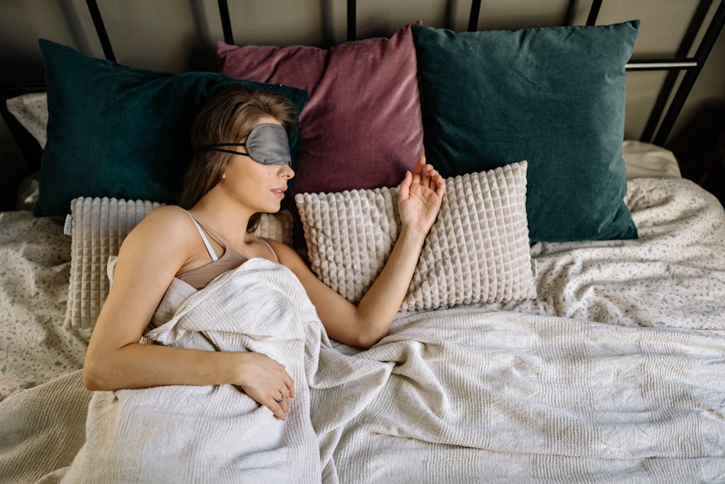 A woman sleeping with an eye mask on a cozy bed, surrounded by pillows and blankets.